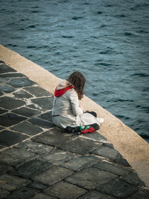 Free A woman in a coat sits pensively on a stone path by the calm sea. Stock Photo