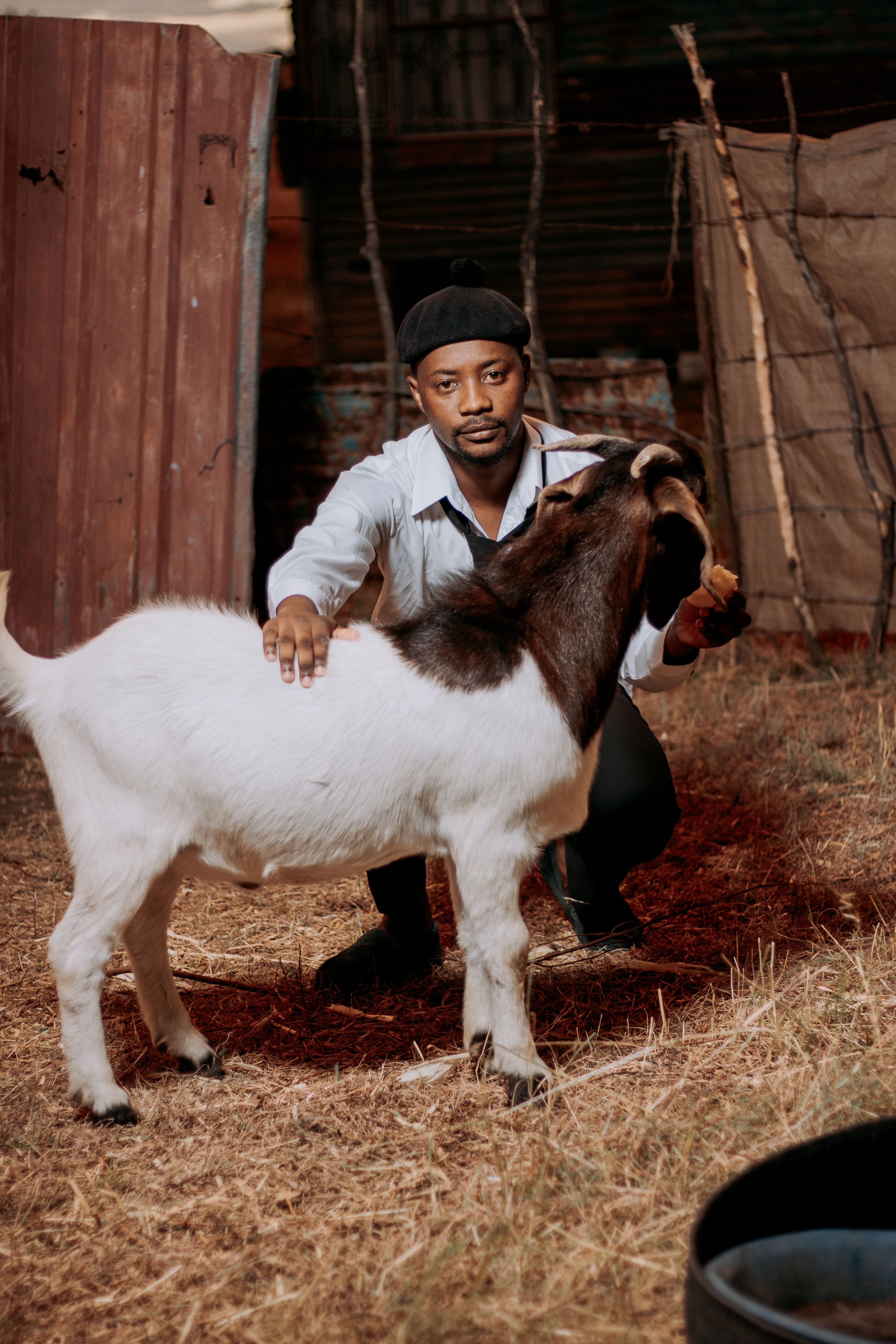 A man with a black hat cares for a white and brown goat outdoors on a farm, showing human-animal connection.