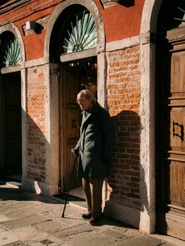 Elderly man with cane strolling past historical Venetian architecture in sunlight.