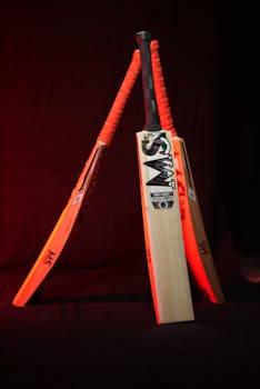 Artistic arrangement of three cricket bats with orange handles and a warm lighting backdrop.