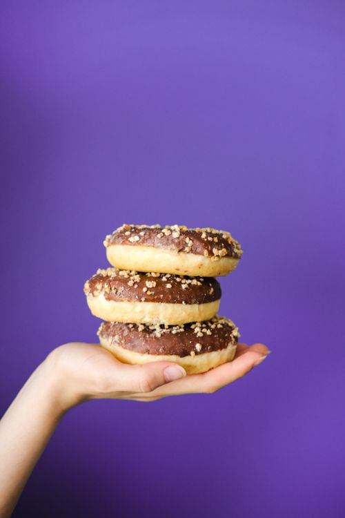 Free A hand holds three stacked chocolate donuts against a vivid purple background. Stock Photo