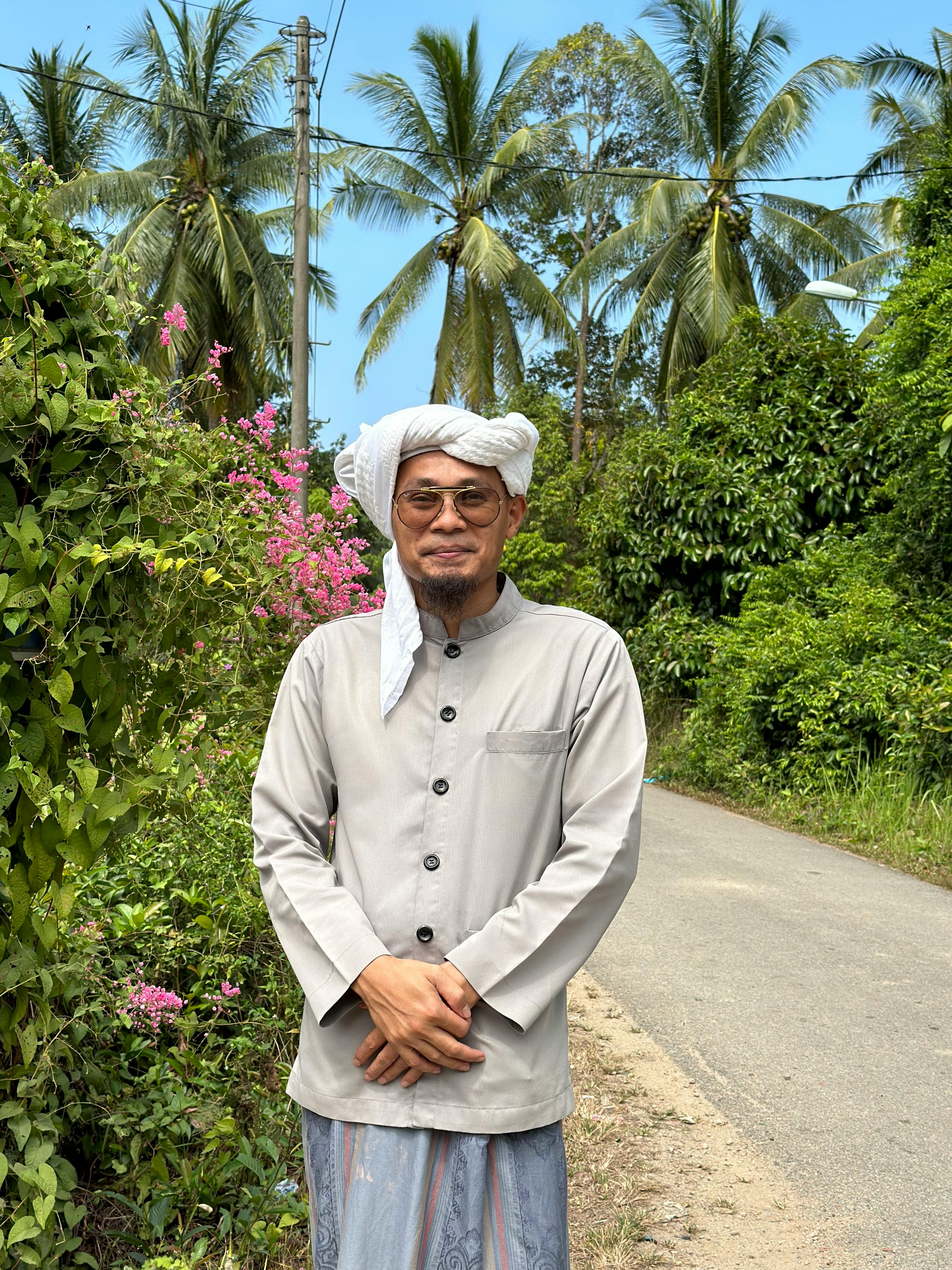 Man in traditional attire stands along a rural Malaysian road surrounded by lush greenery and palm trees.