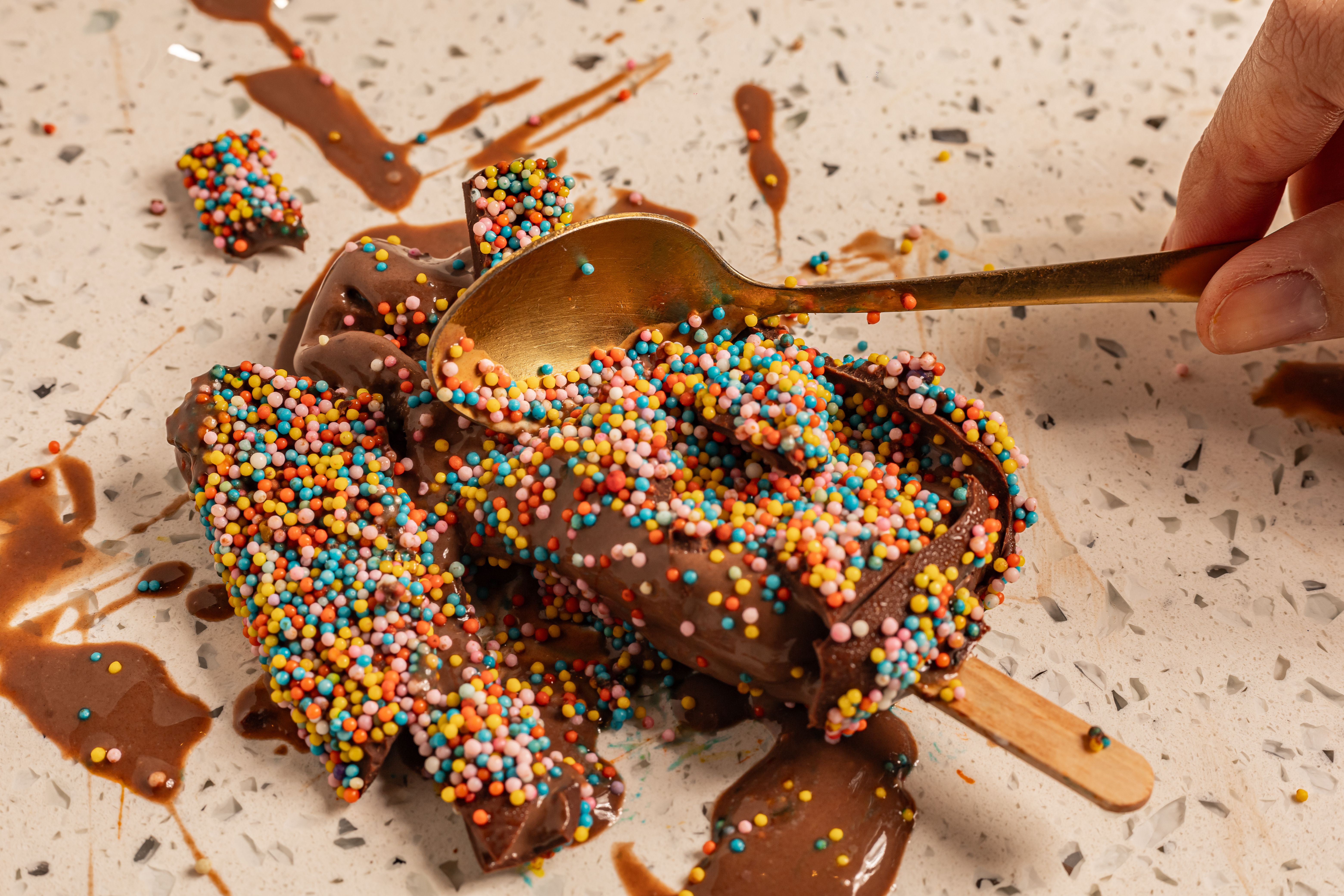 A frozen banana dipped in chocolate and rolled in rainbow sprinkles, held against a bright yellow background