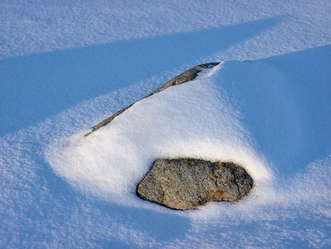 A single rock partially covered in fresh snow, casting a shadow in the winter sunlight.