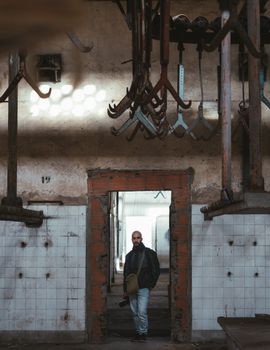 A man stands in a doorway of an abandoned industrial building surrounded by hanging hooks.