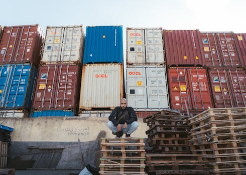A man crouches on wooden pallets in front of colorful stacked shipping containers, outdoor scene.