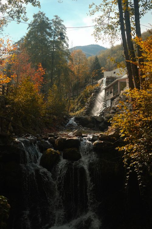 Free A serene waterfall flows through a colorful autumn forest with mountains in the background. Stock Photo