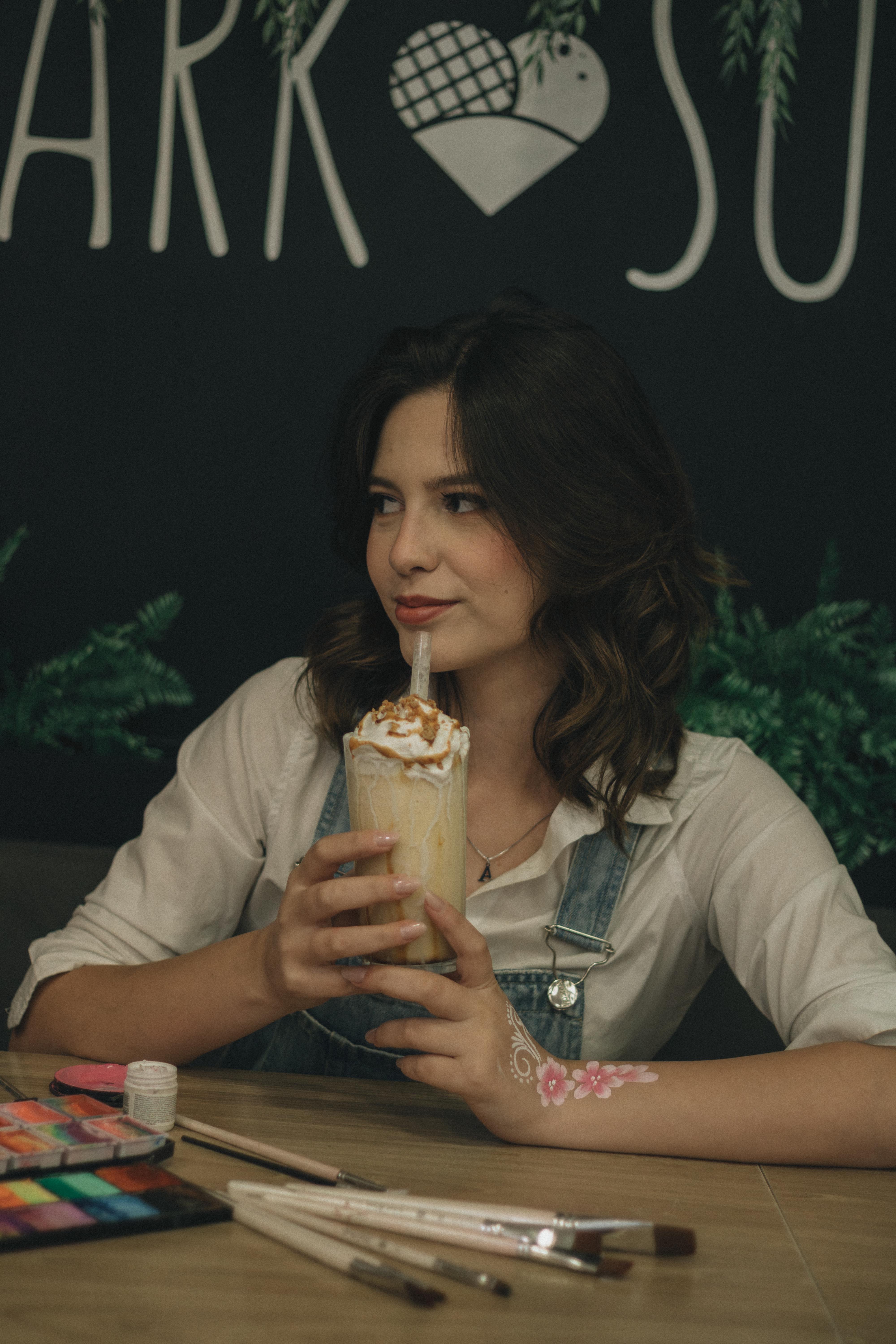 Безкоштовне Молода жінка сидить за столиком у кафе, насолоджуючись молочним коктейлем, оточена художнім приладдям. стокове фото