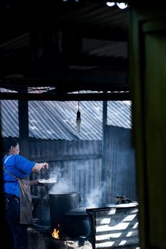 Woman cooking over open fire in an outdoor kitchen, using large pots.
