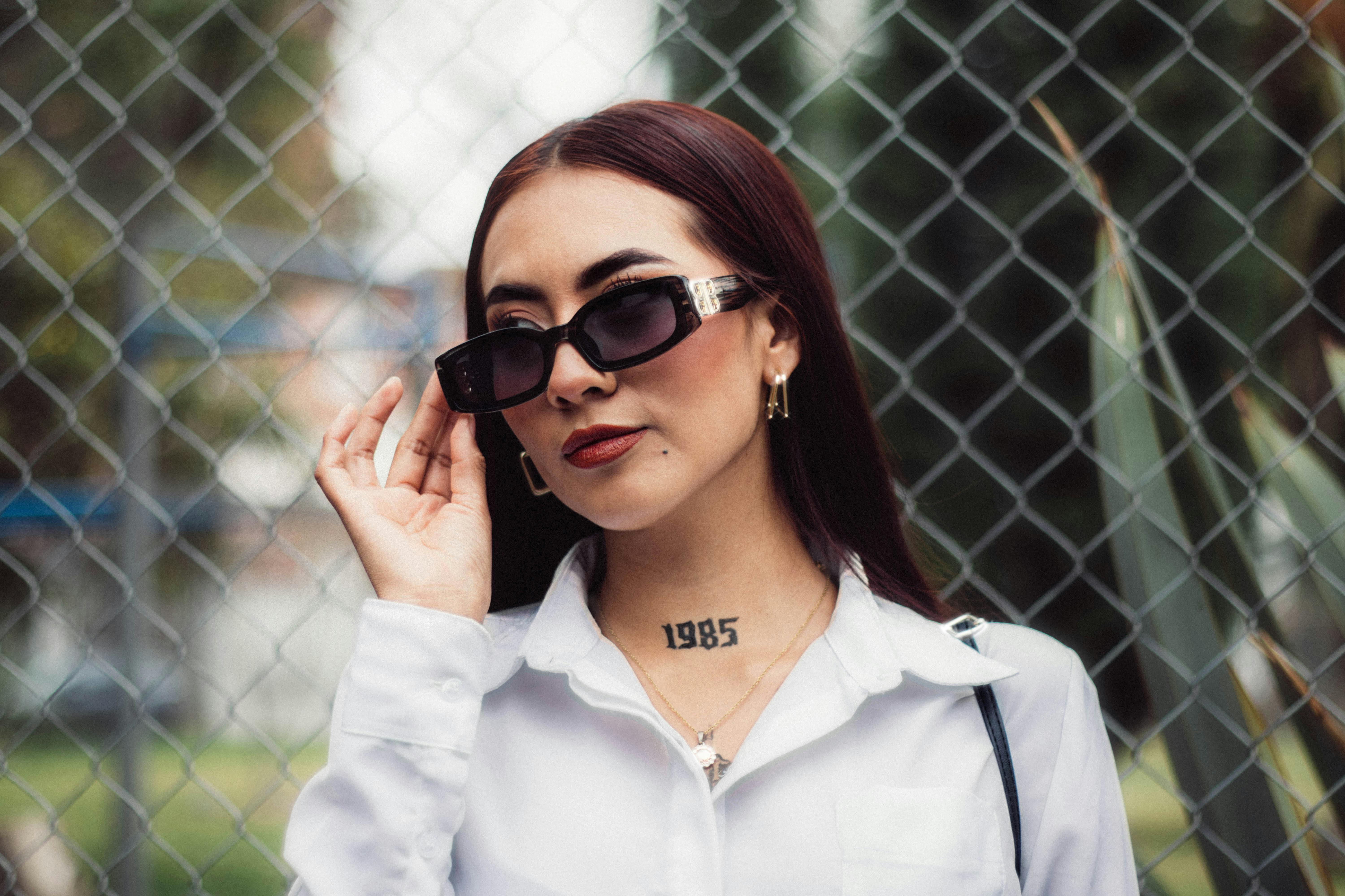 ฟรี หญิงสาวท่าทางมั่นใจ สวมแว่นกันแดดและเสื้อเชิ้ตสีขาว โพสท่าอยู่ข้างรั้ว คลังภาพถ่าย