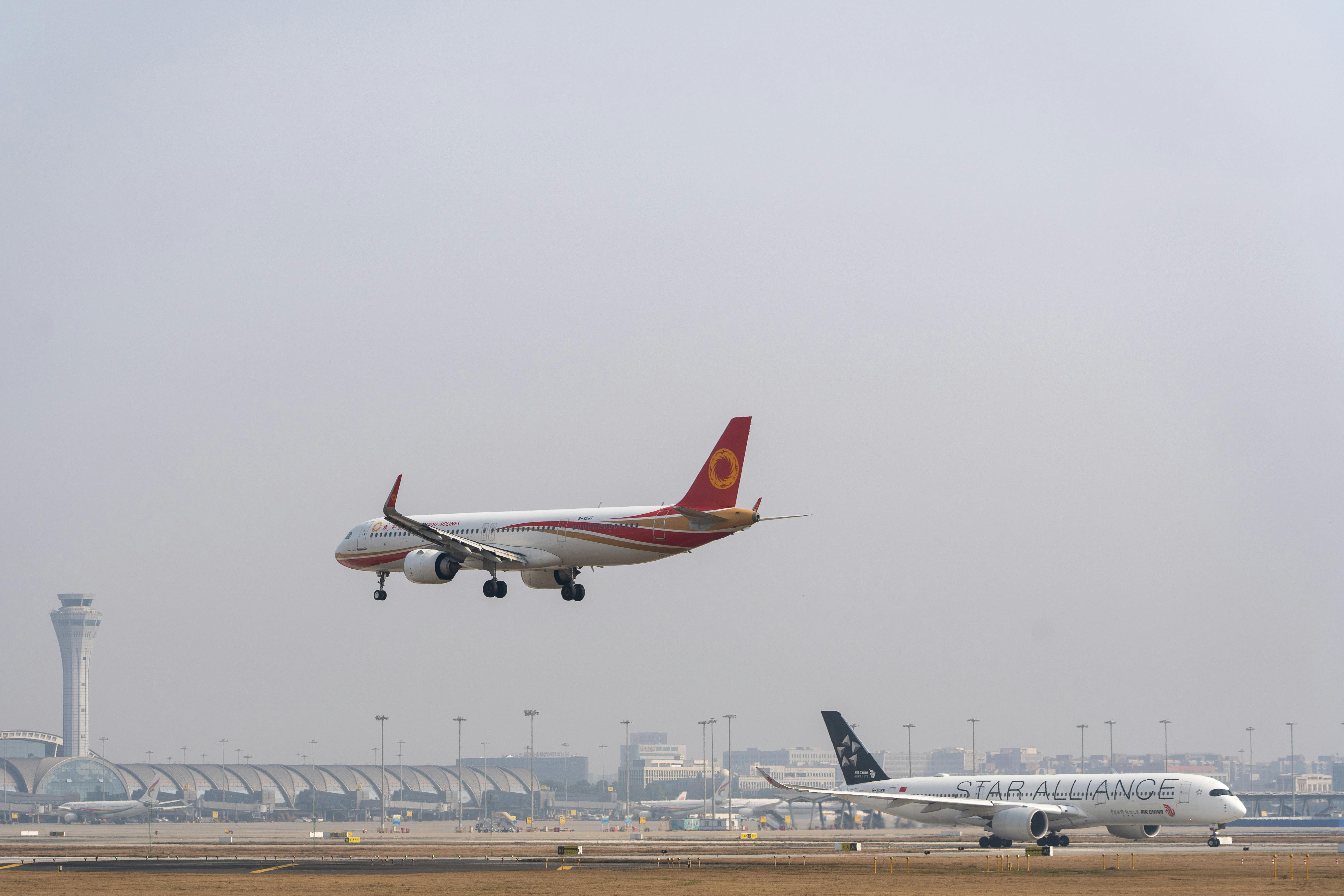 Two commercial airplanes approaching landing at a busy international airport with control tower visible.