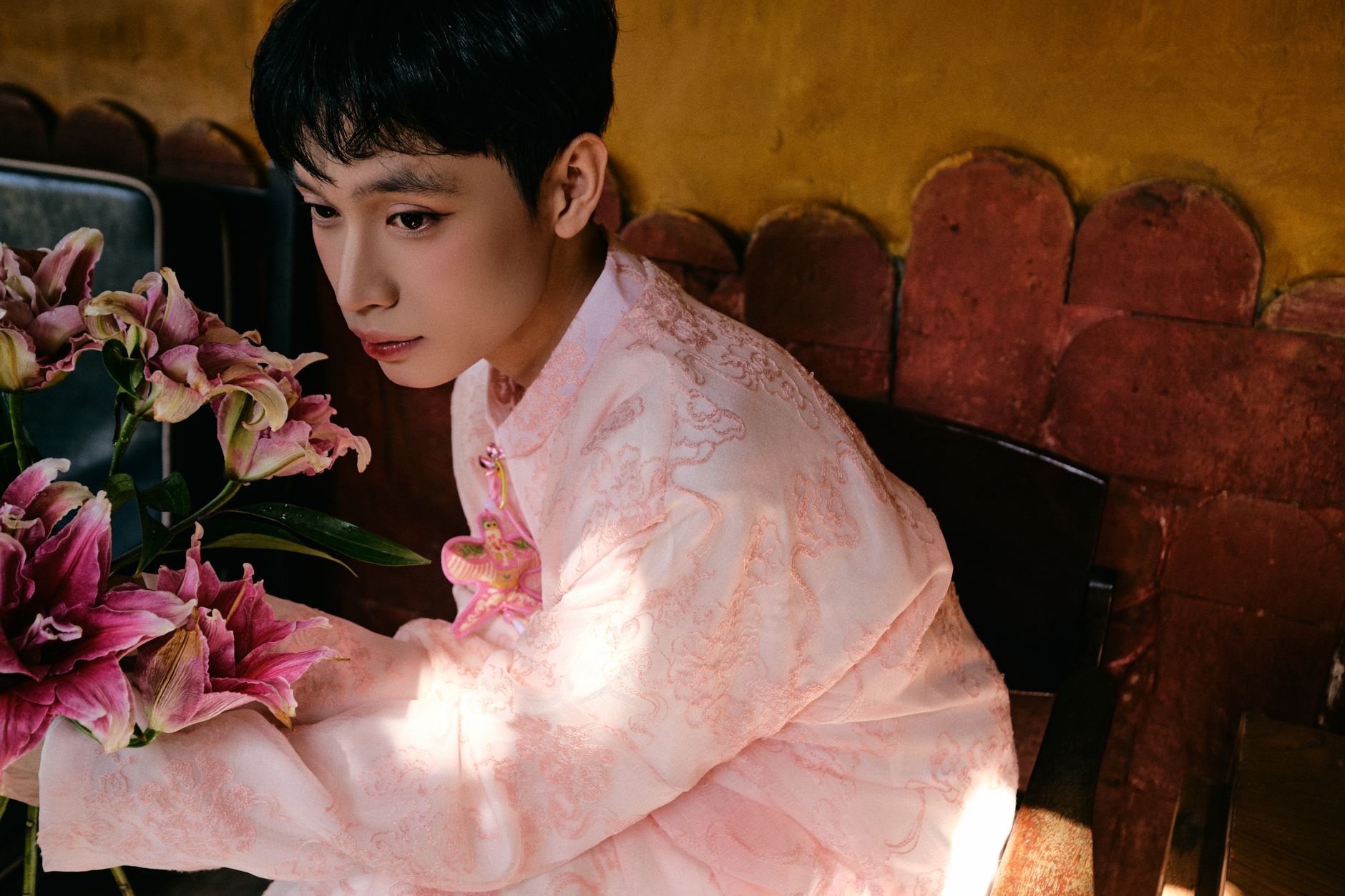 A man in a pink Vietnamese Ao Dai holding flowers indoors.