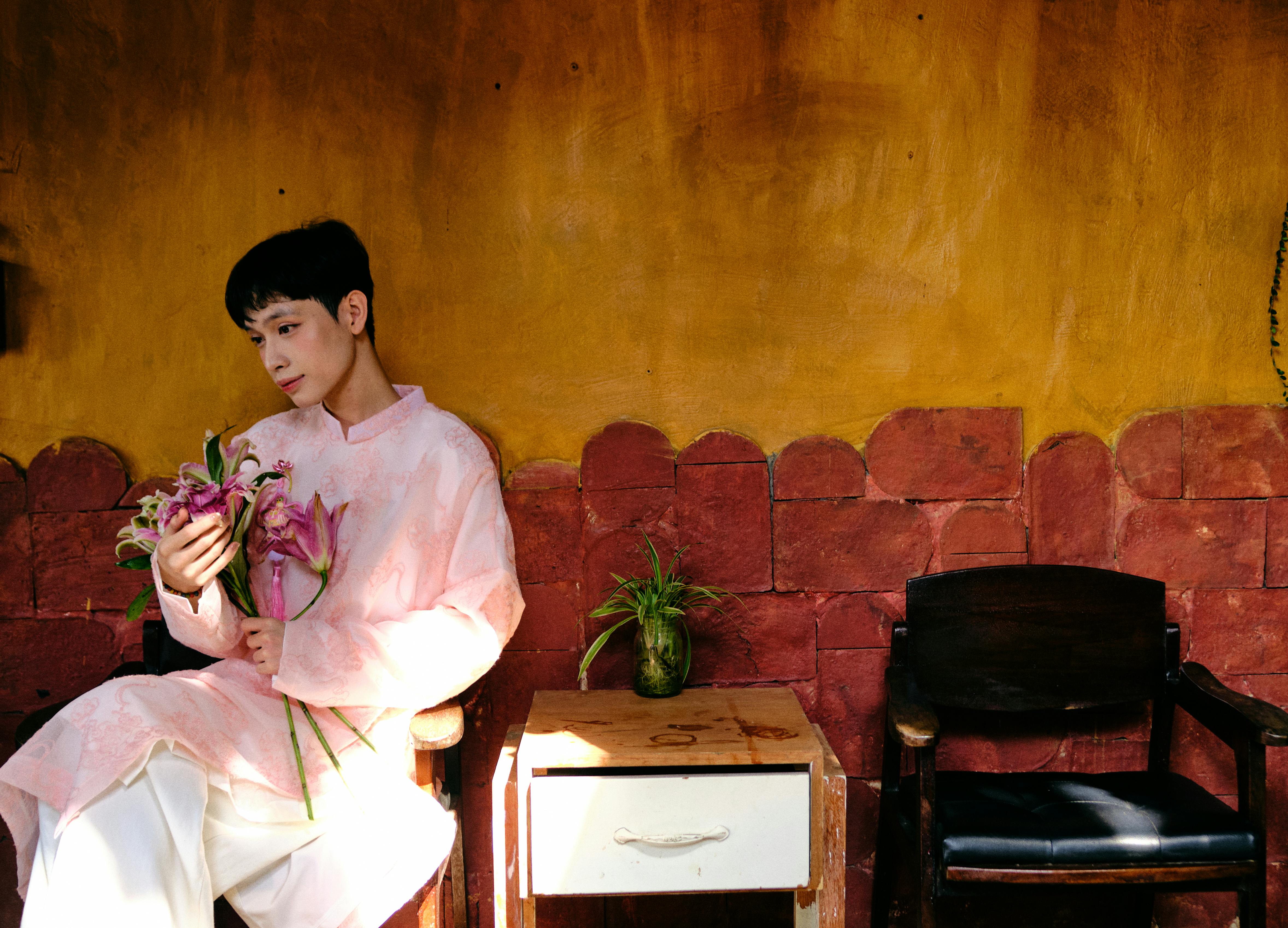 Young man in traditional pink Ao Dai holding lilies, seated indoors in Vietnam.