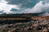 Grass area covered with stones against hills with clouds touching peaks in overcast sky