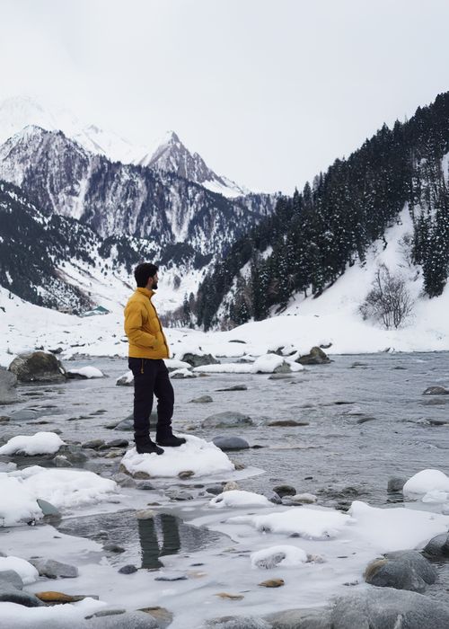 Free Man in a yellow jacket stands by a snowy river amidst mountainous landscape, capturing winter serenity. Stock Photo