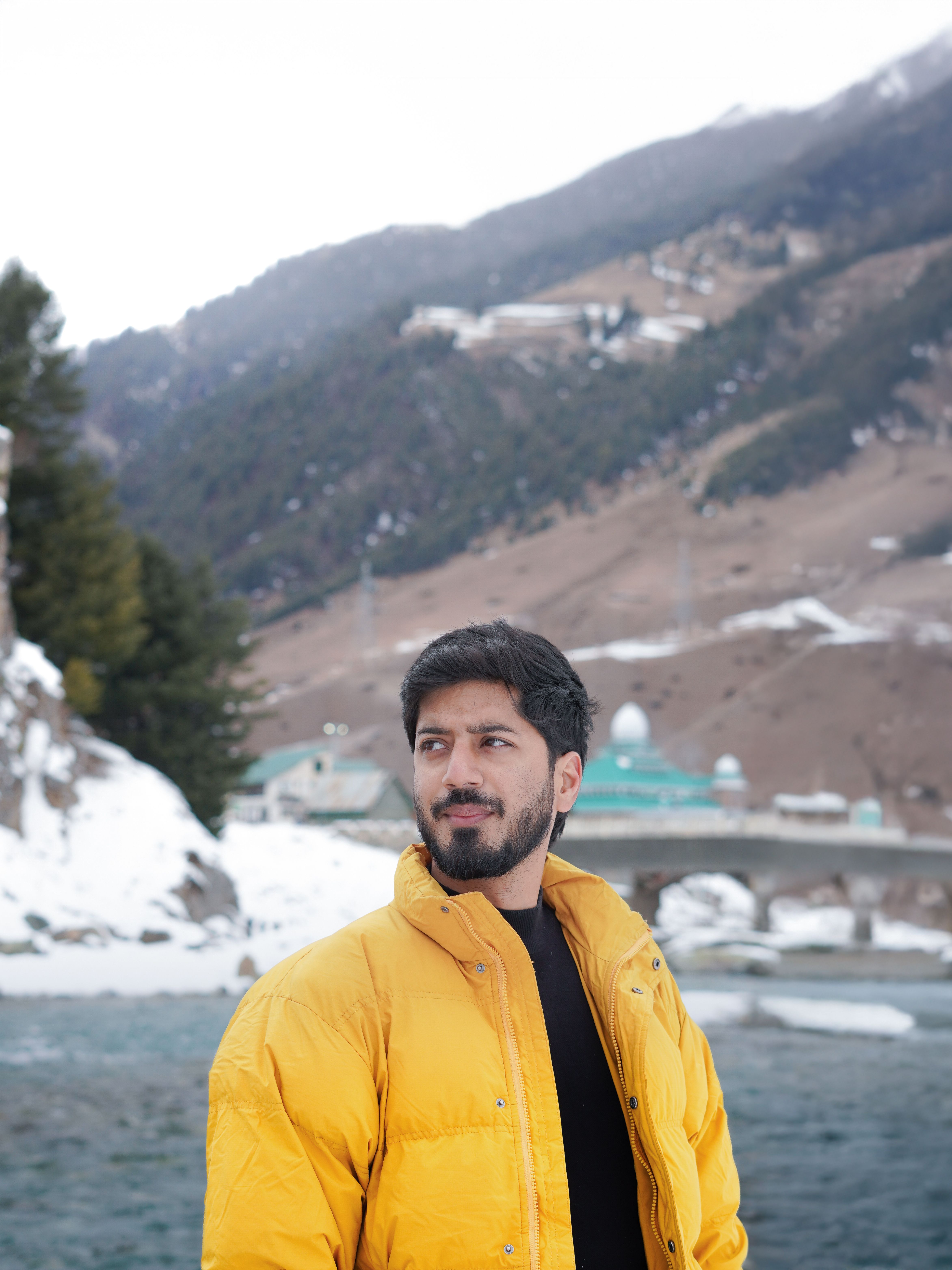 Man in a bright yellow jacket stands before a snowy mountainous landscape by a bridge.