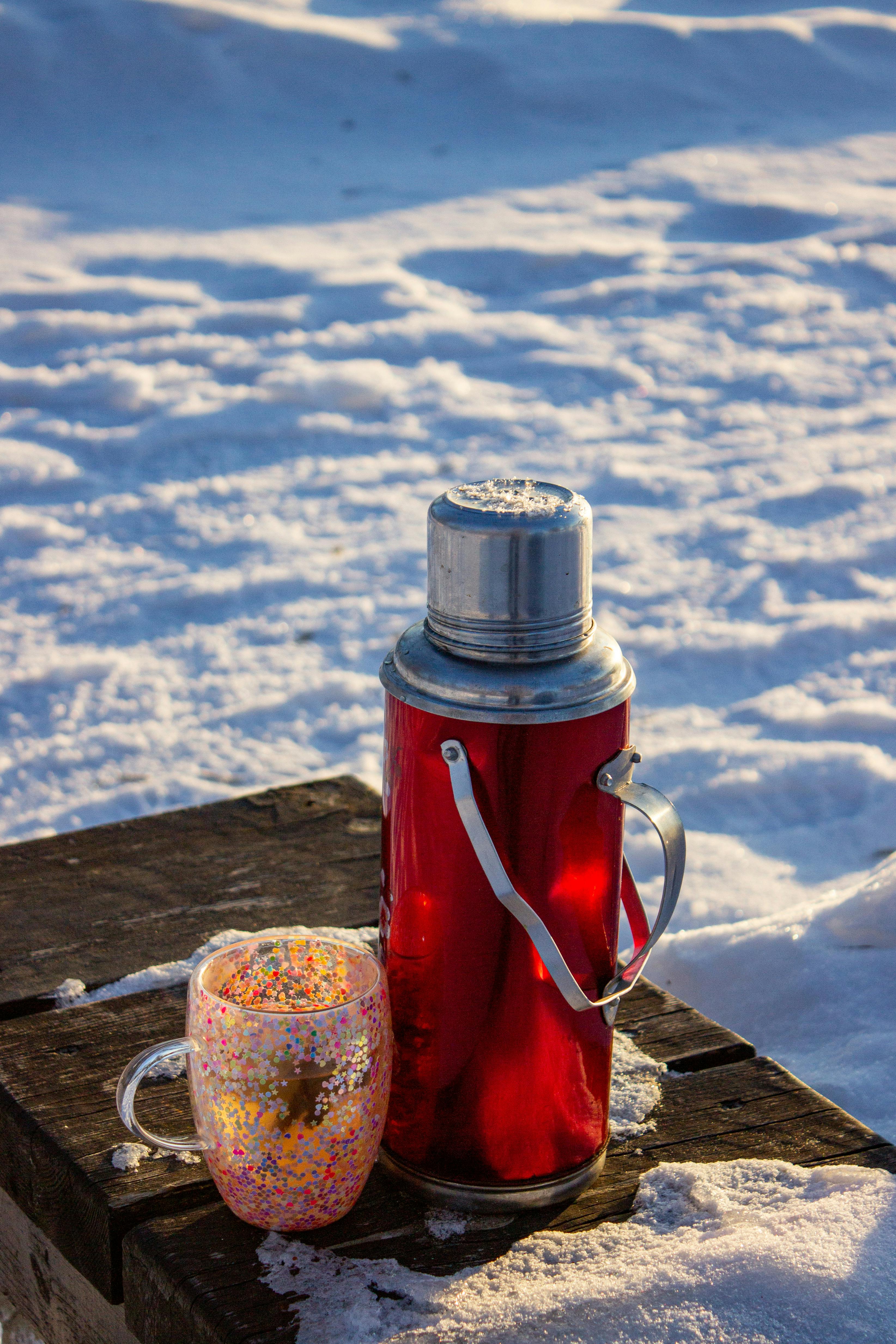 A red thermos and colorful mug on a snowy wooden bench, capturing a winter outdoor setting.