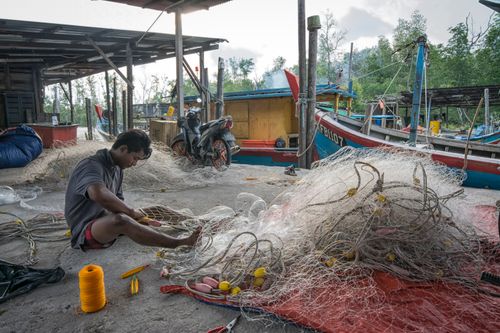 Free Man mends fishing nets by the harbor, surrounded by boats and fishing equipment. Stock Photo