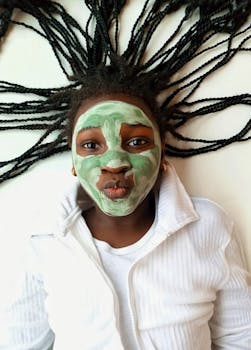 Smiling girl with braided hair and green facial mask, enjoying spa day indoors.