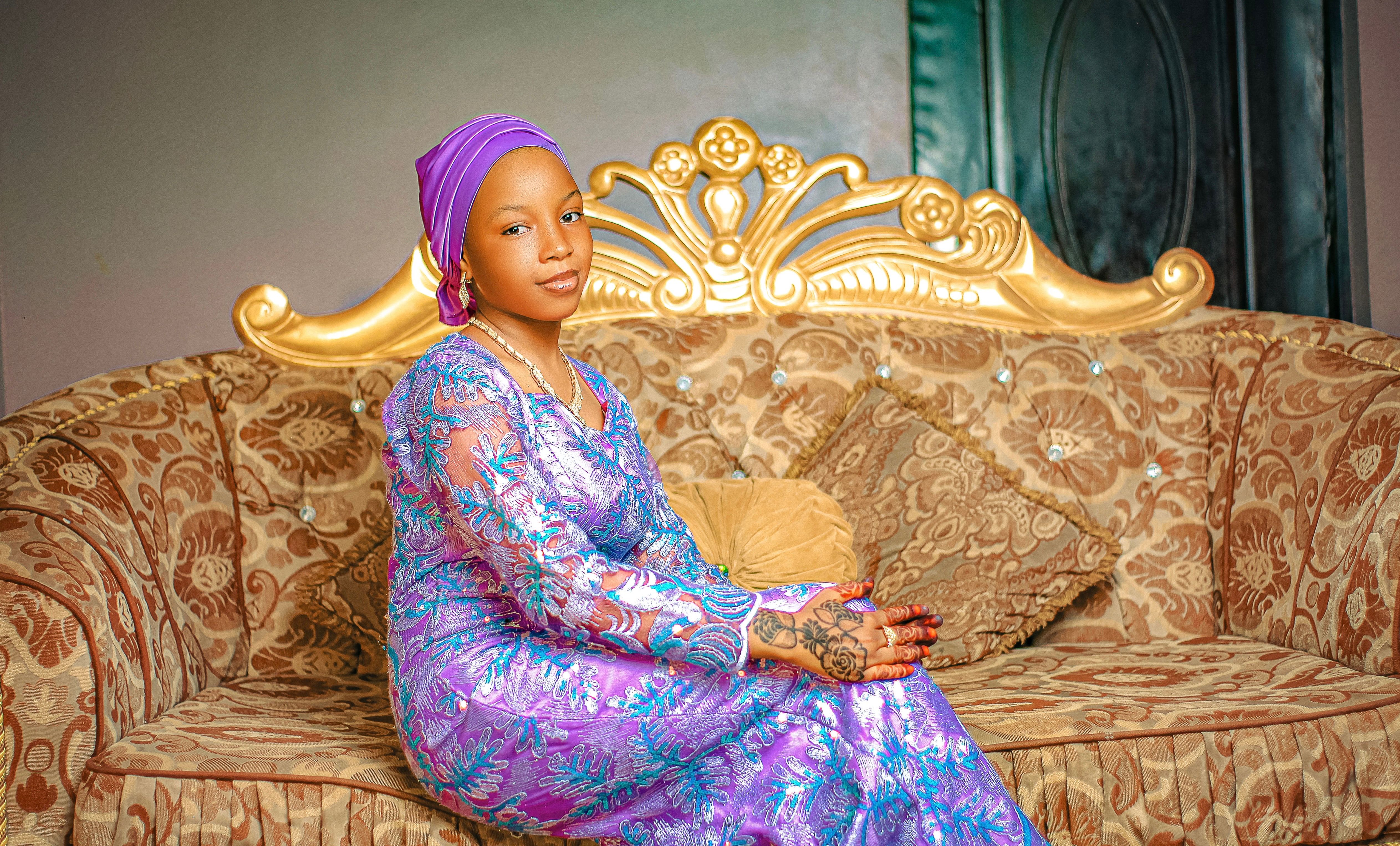 A woman in vibrant traditional attire sits elegantly indoors on a patterned sofa.