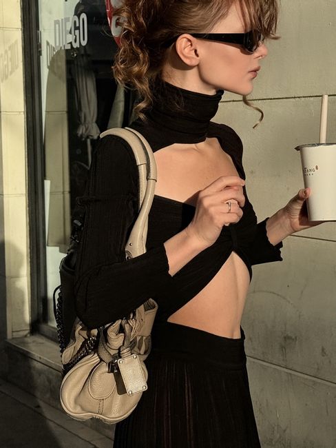 Stylish woman in trendy outfit holding a drink in urban sunlight.