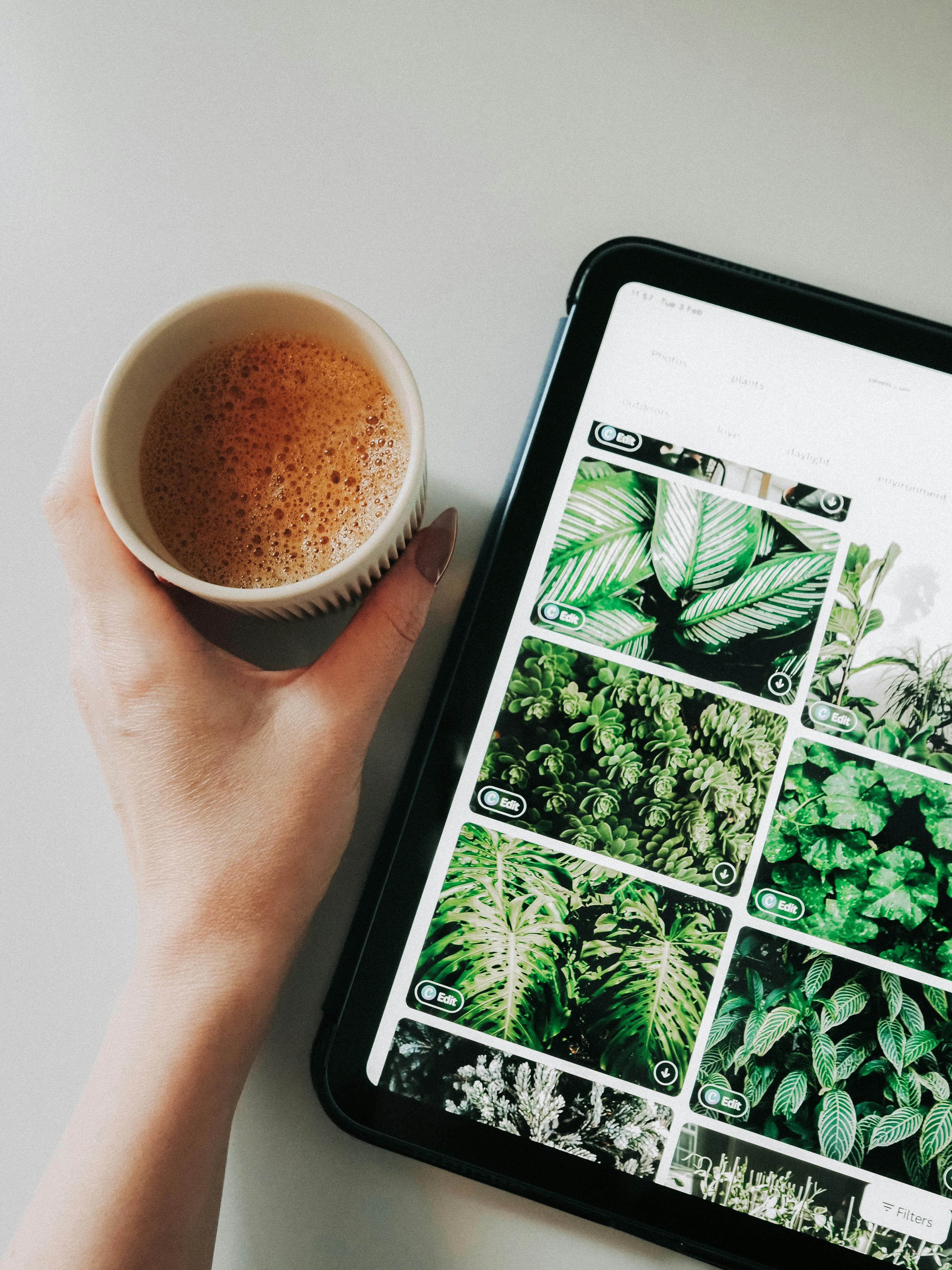 Hand holding coffee cup beside a tablet displaying vibrant plant images, showcasing a modern digital lifestyle.