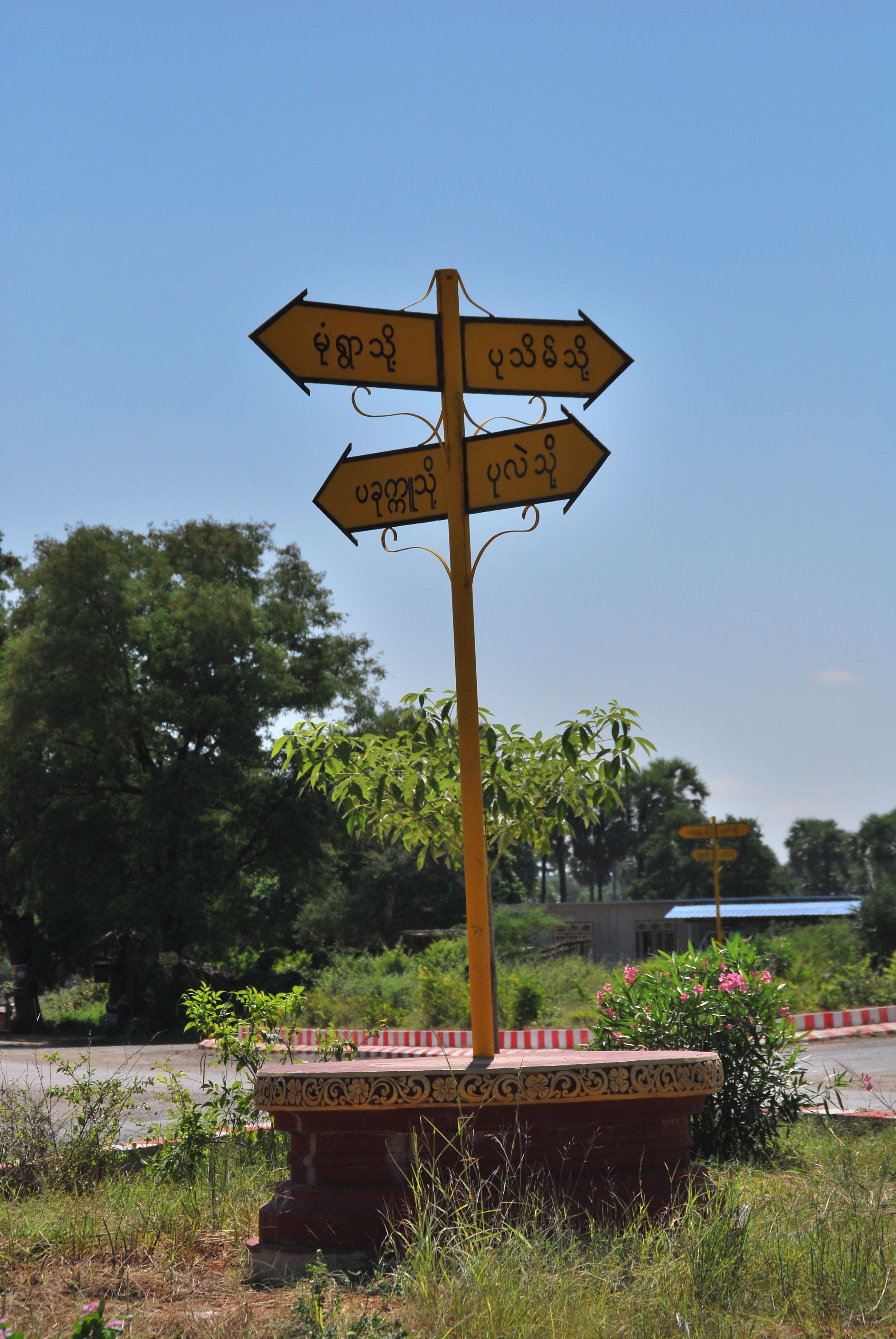 A yellow directional signpost with Myanmar script set against a blue sky and lush greenery.