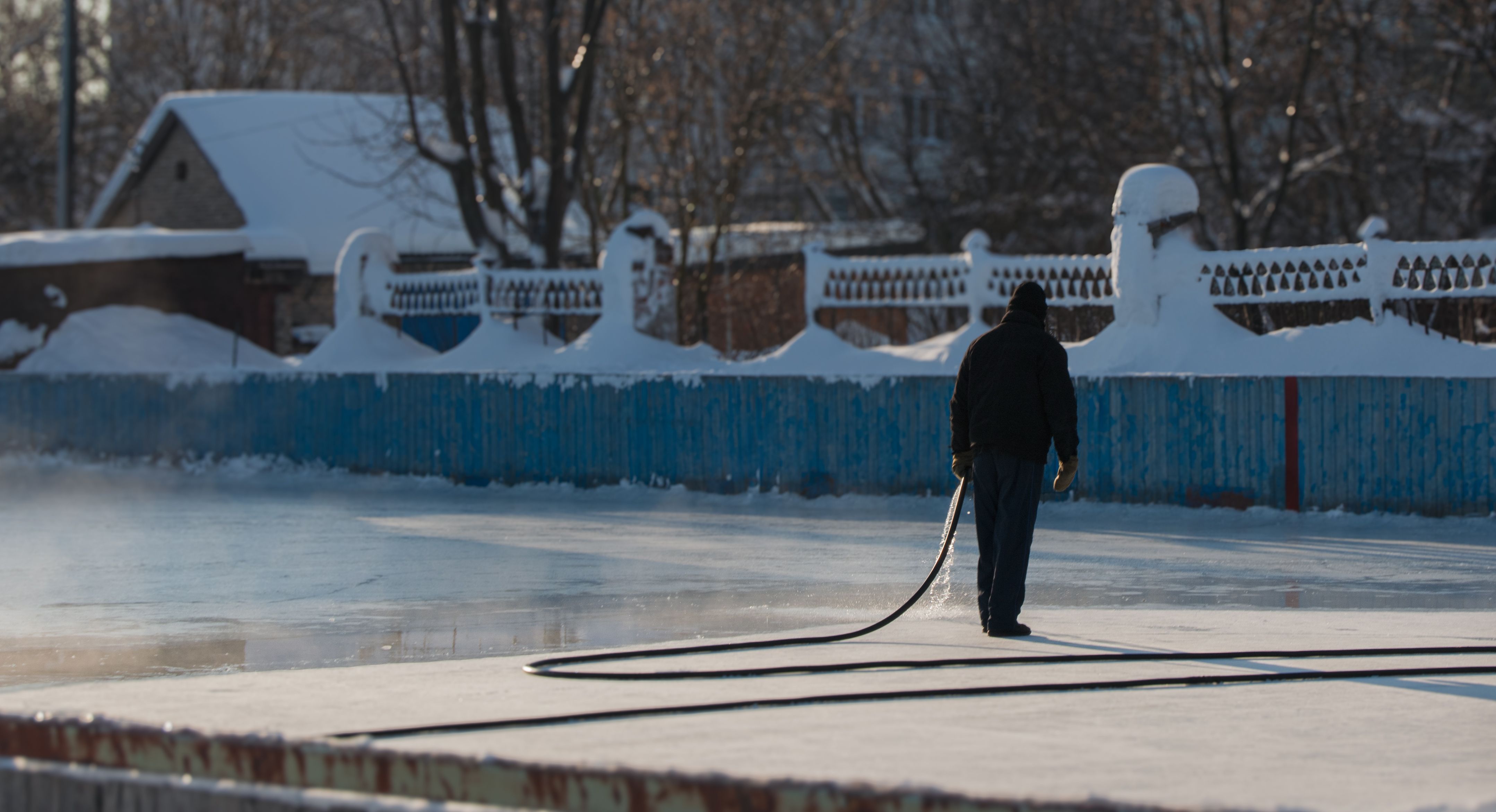 A person using a hose to flood and maintain a homemade outdoor ice rink in winter, illustrative of a backyard rink setup