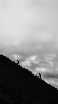 Two silhouettes climbing a steep hill under a dramatic cloudy sky in black and white.