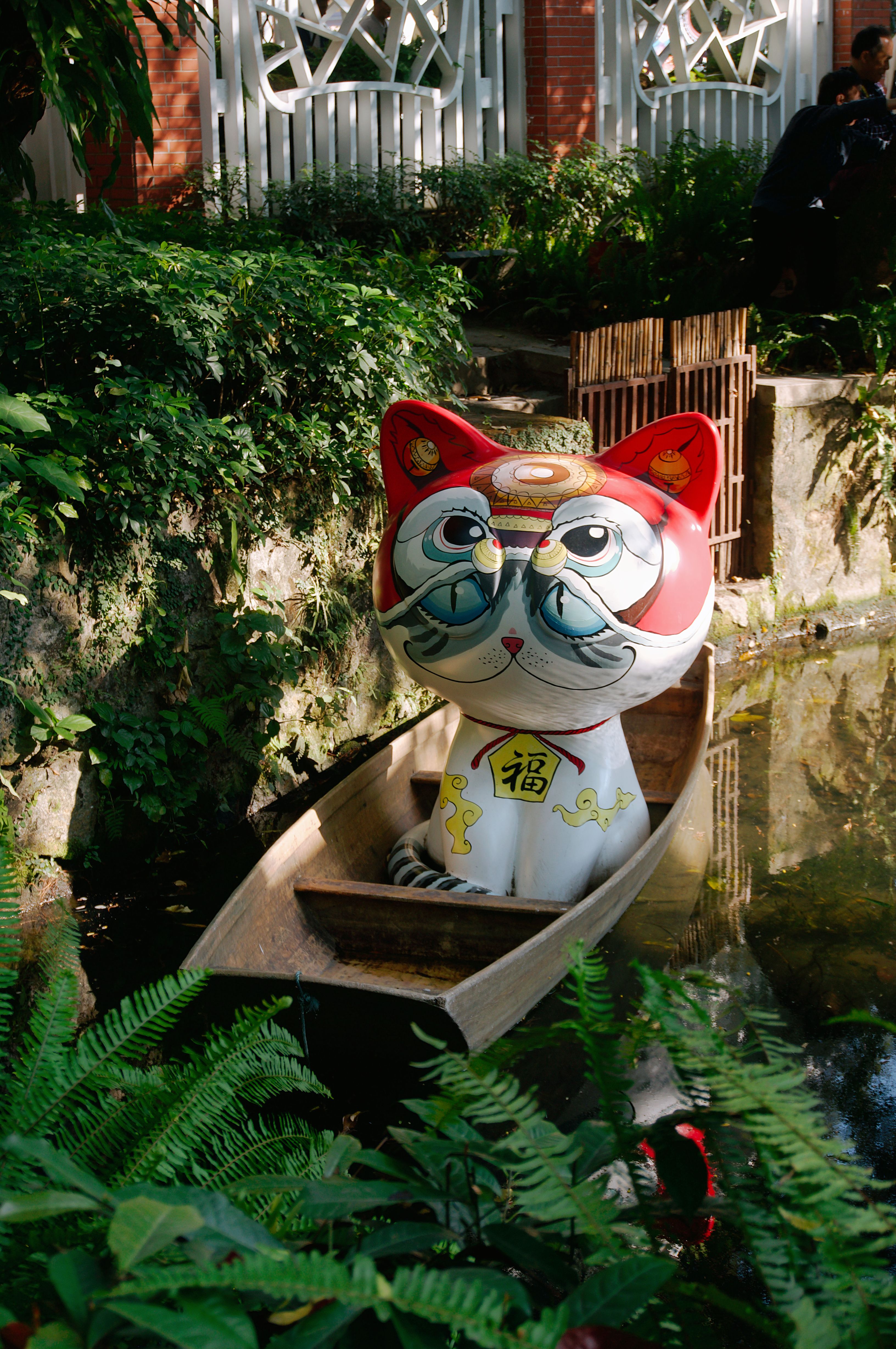 A vibrant Maneki-Neko statue sits in a canoe surrounded by lush greenery and water.
