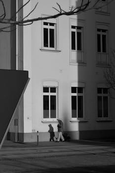 Black and white photo capturing a building facade with stark shadow play and a passing figure.