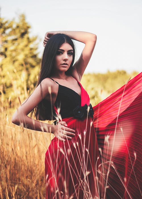 Gorgeous young ethnic female with long dark hair in elegant dress standing in dry tall grass with hand on waist and looking at camera on sunny day