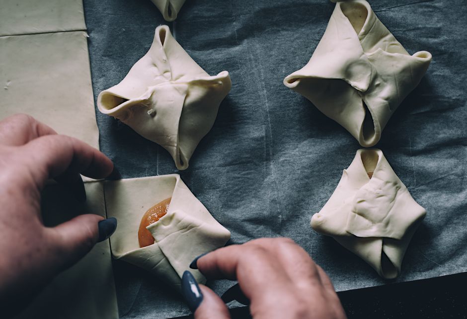 Close-up of hands folding pastry parcels on parchment paper in a kitchen setting.