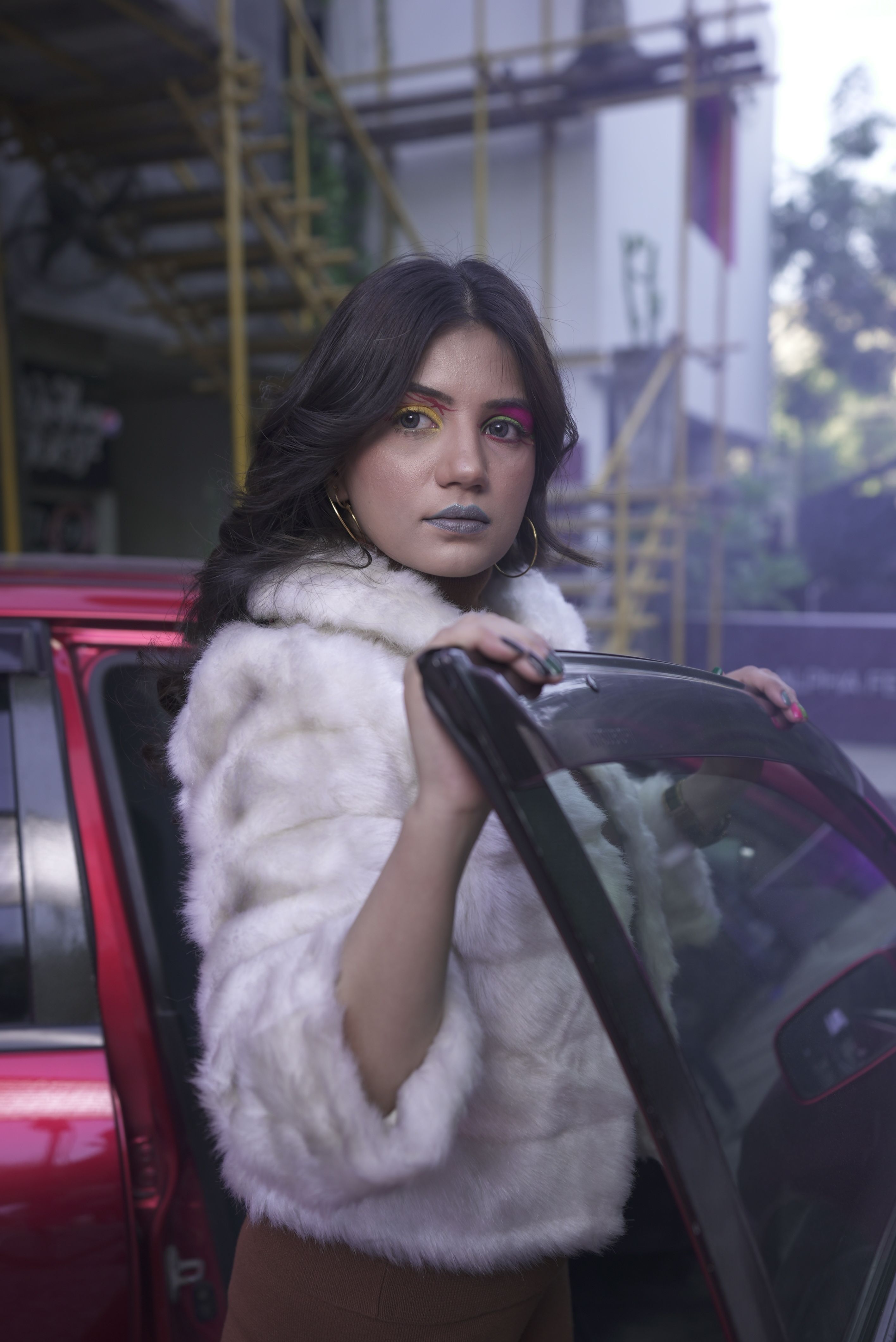 A stylish woman with bold makeup and a faux fur coat steps out of a red car, showcasing modern fashion.