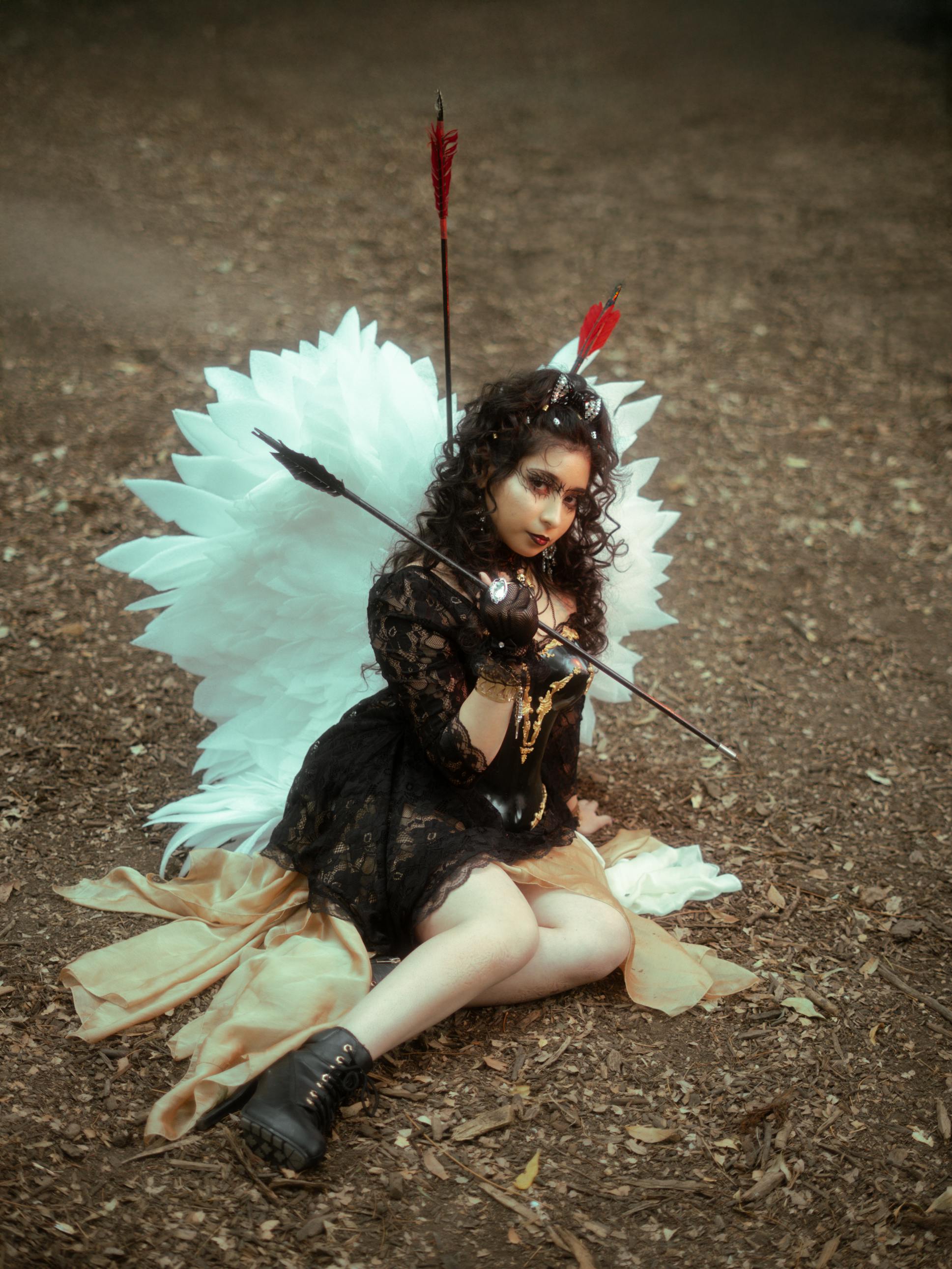 Gratis Una mujer vestida como un ángel de fantasía con alas y flechas, sentada en el suelo de un bosque. Foto de stock