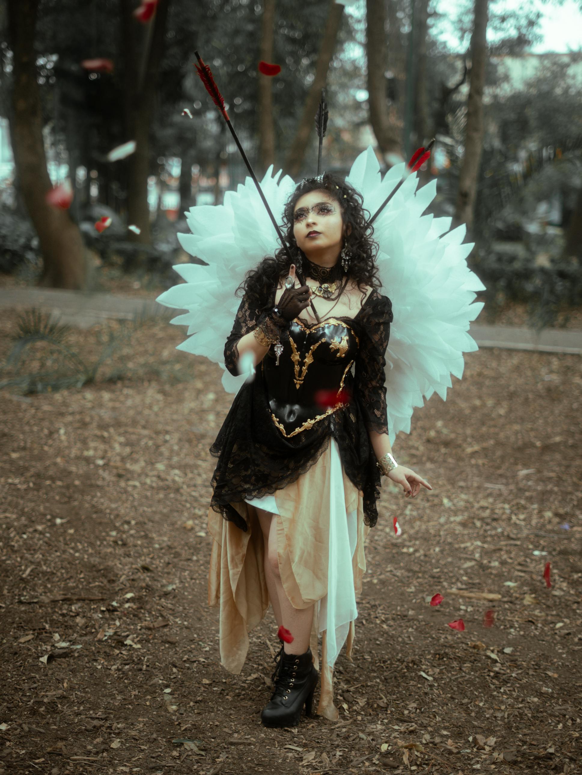 Gratis Seorang wanita dengan kostum bak malaikat dan bergaya gotik berdiri di hutan yang dikelilingi oleh kelopak bunga yang berguguran. Foto Stok