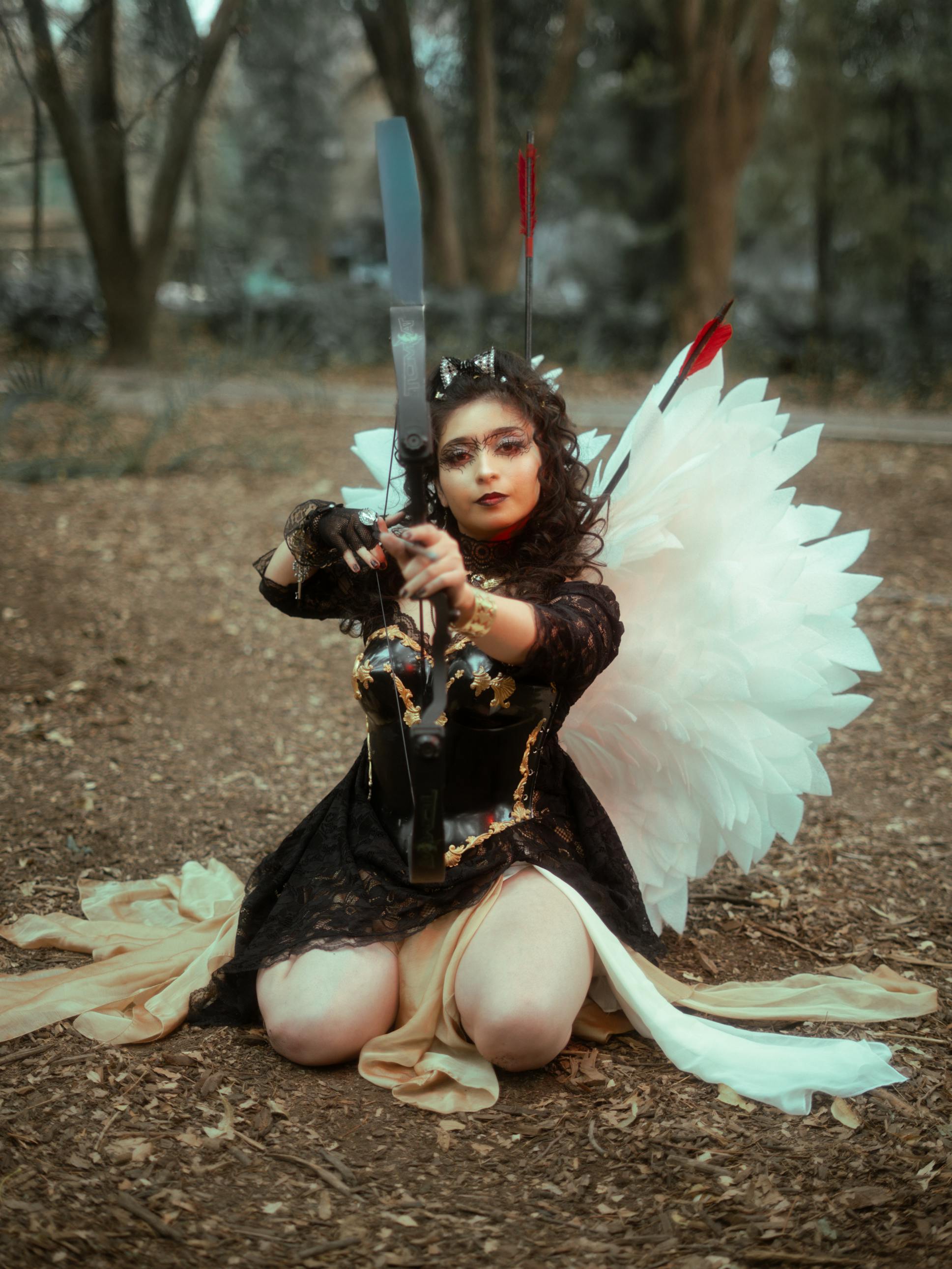grátis Uma mulher vestida de anjo, com temática de fantasia, mirando com um arco em uma floresta. Foto profissional