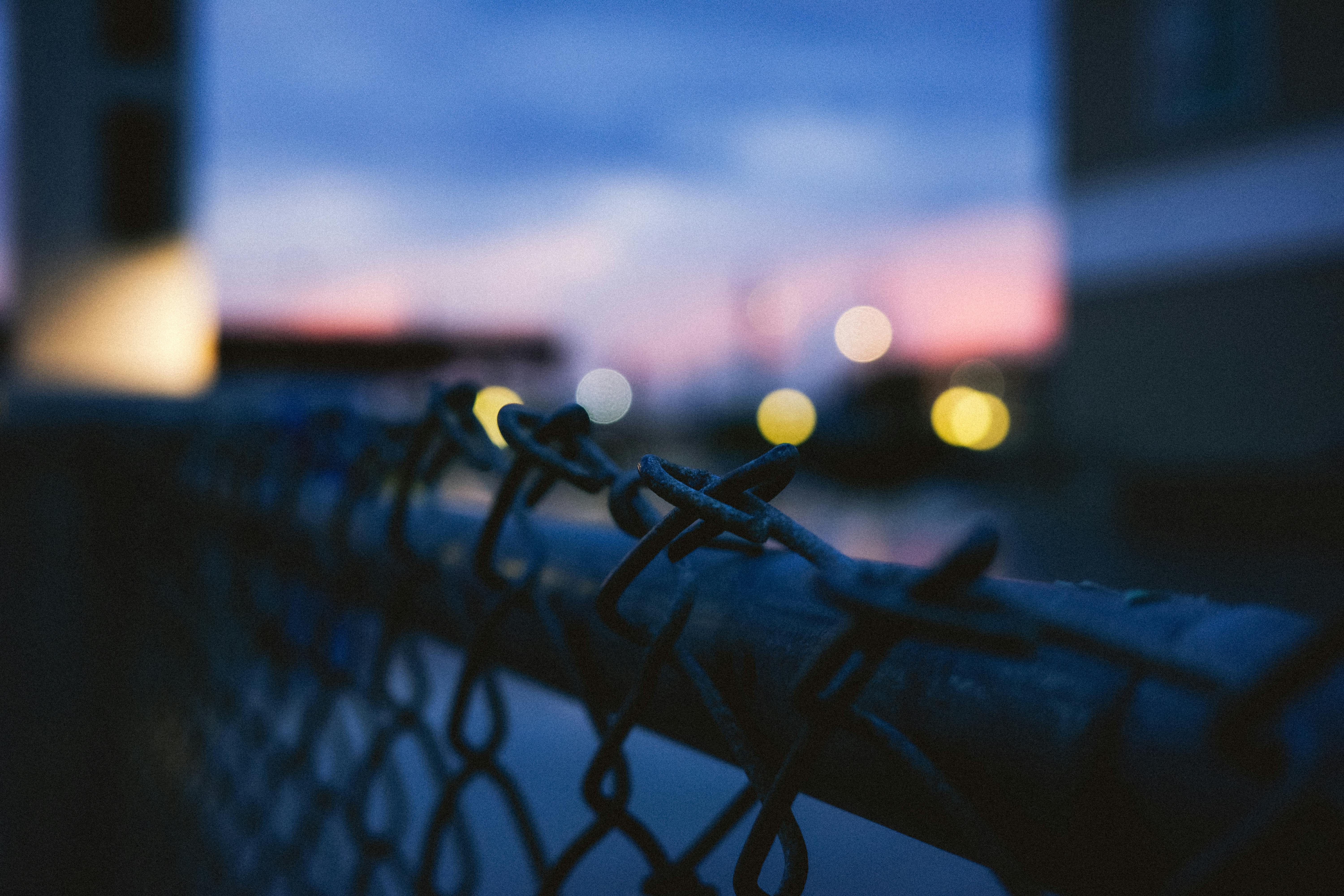 Gratis Gambar close-up pagar kawat dengan lampu bokeh di latar belakang saat senja. Foto Stok