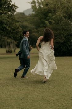 A couple in formal attire joyfully runs across a grassy field during daytime, surrounded by lush greenery.