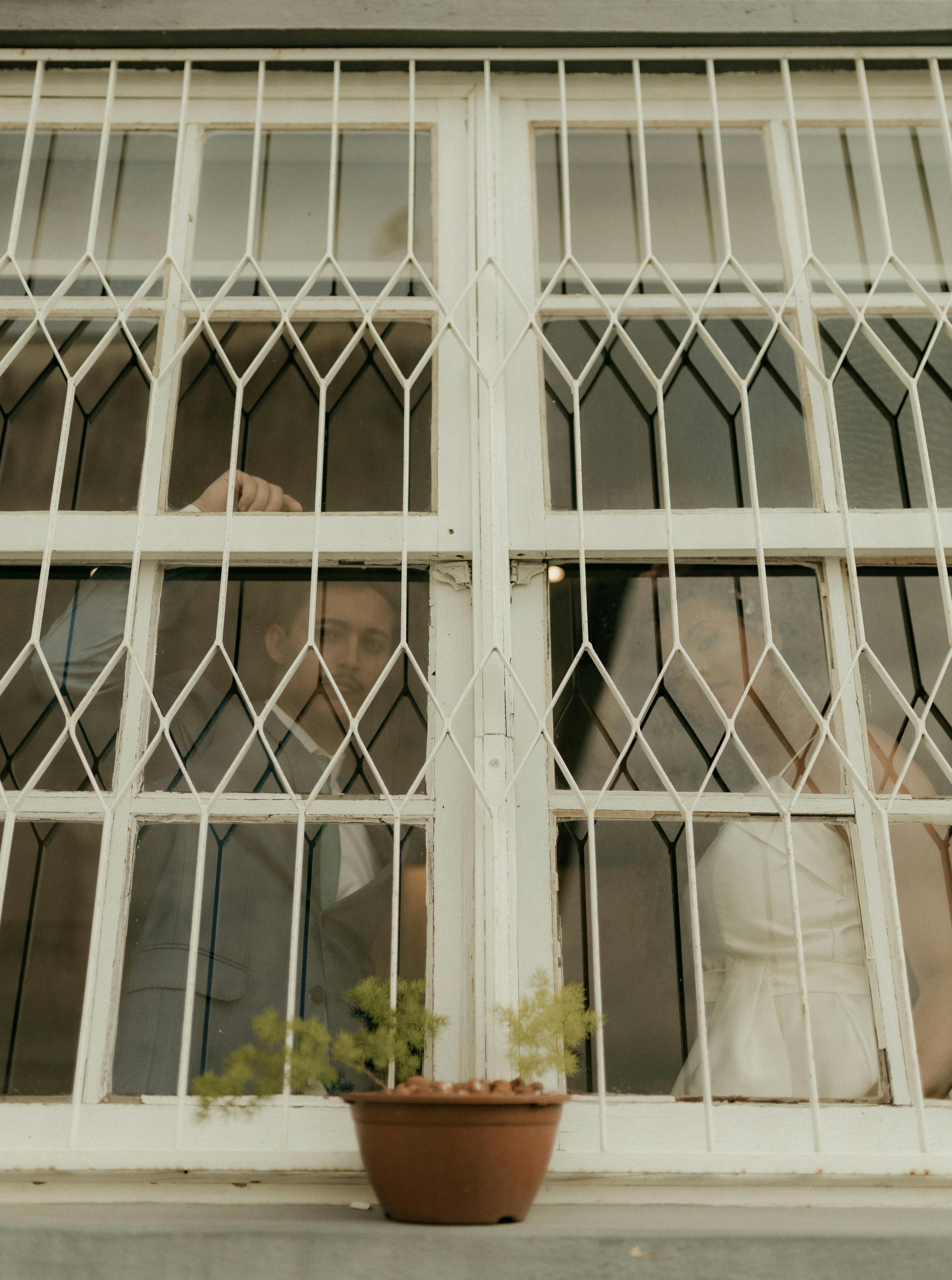 Gratis Una pareja mirando a través de una ventana vintage con un patrón de cuadrícula blanca, creando una escena interior íntima. Foto de stock