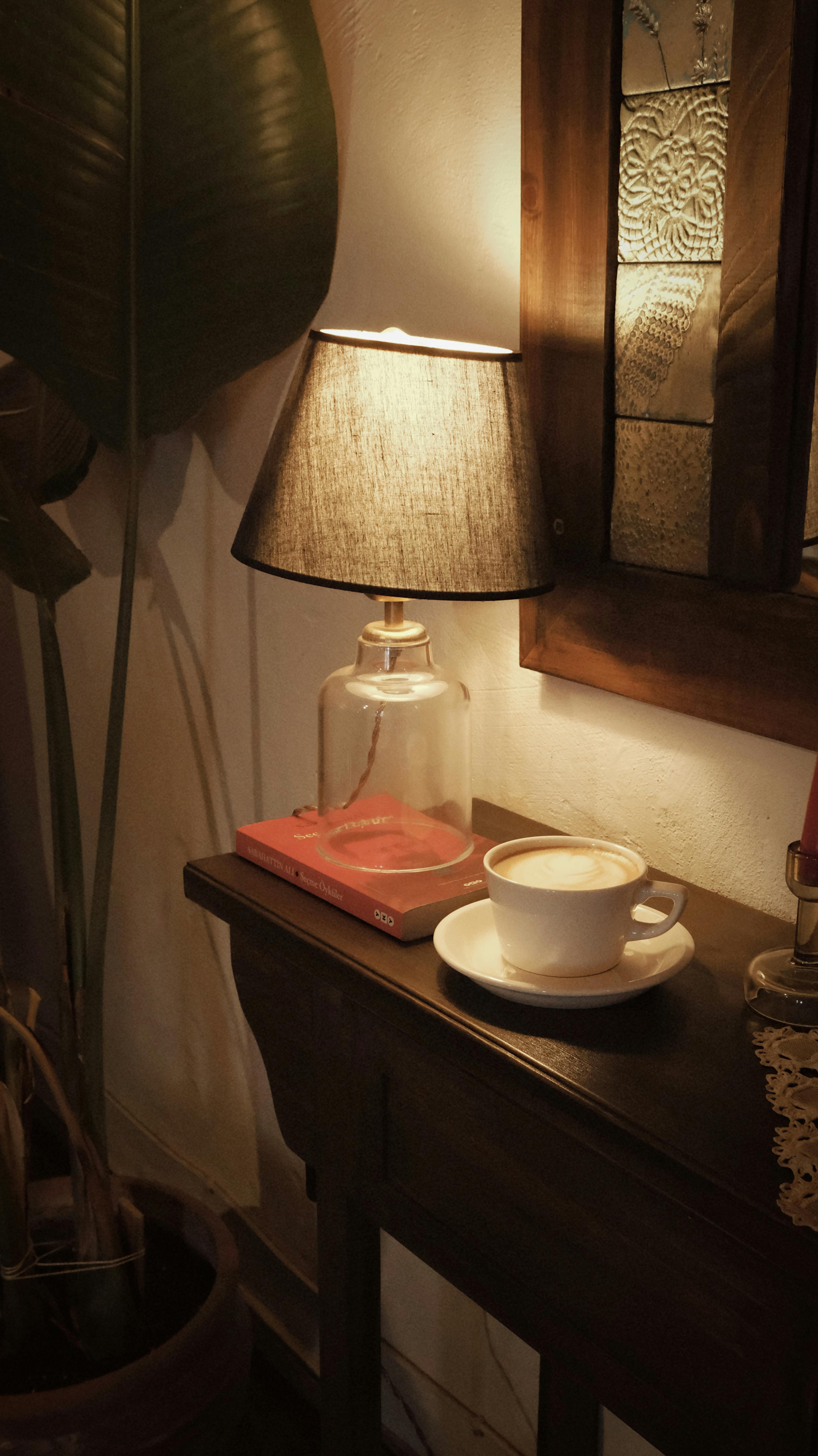 grátis Ambiente interno aconchegante com um abajur, um livro e uma xícara de café sobre uma mesa de madeira. Foto profissional