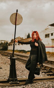 Red-haired woman poses fashionably on train tracks with industrial backdrop.