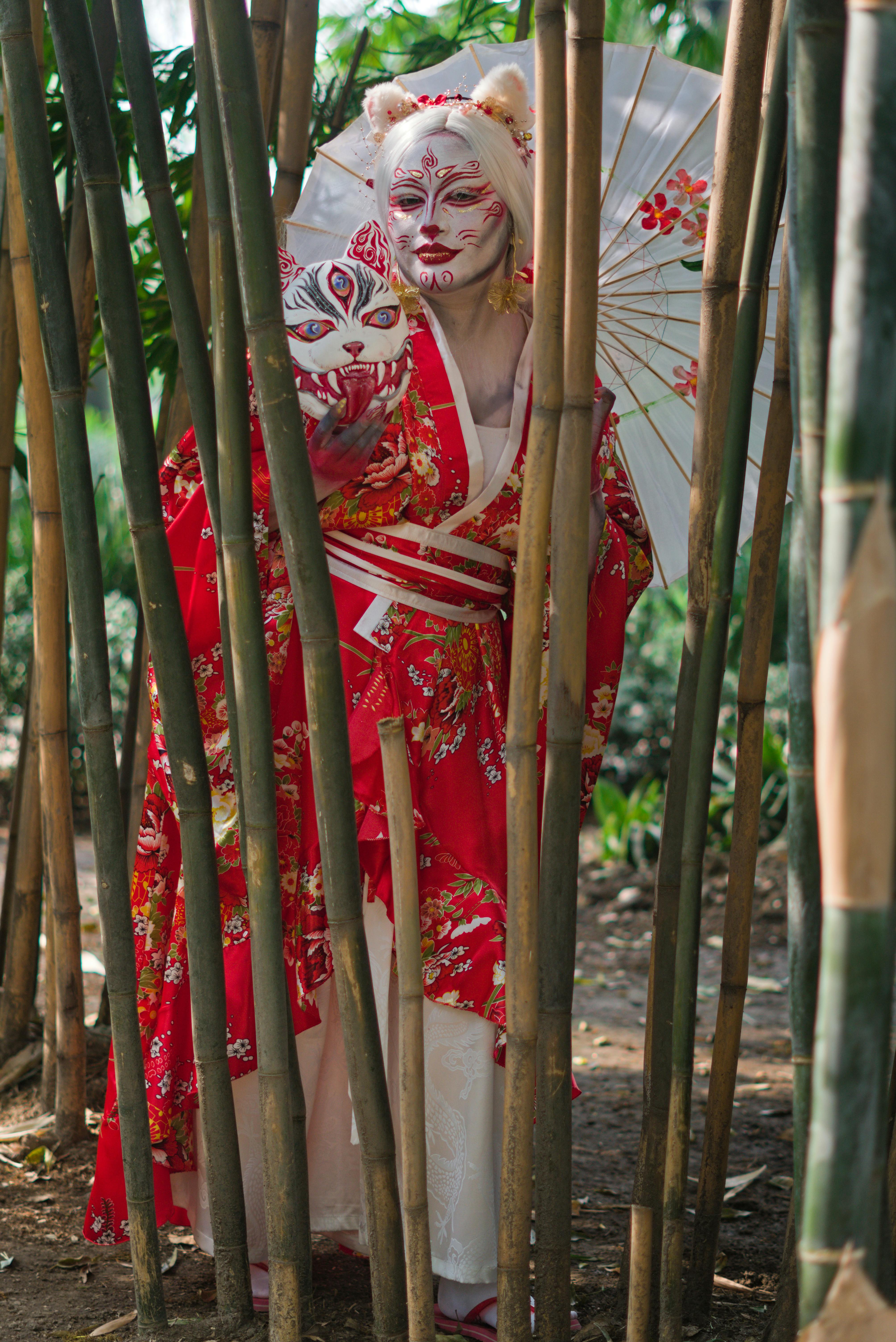 Gratis En geisha i rød kimono og maske står midt i en bambusskov og holder en parasol. Lagerfoto