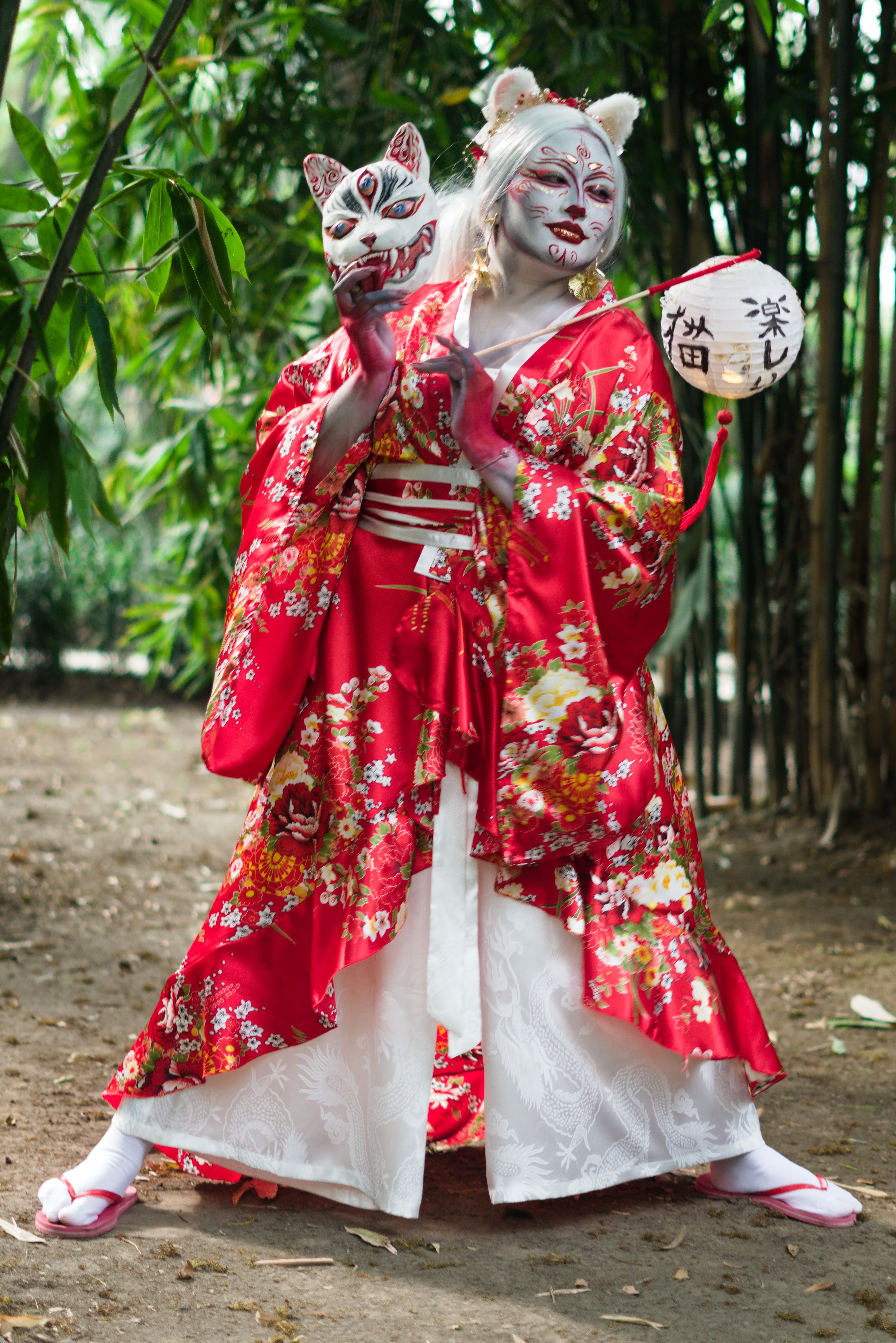 Person in a vibrant red kimono and kitsune mask posing amidst bamboo outdoors.