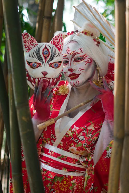Free Artistic portrait of a mystical geisha in traditional red kimono with creative face paint, holding a mythical mask in a bamboo forest. Stock Photo