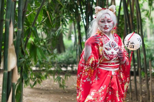 Free Person in vibrant kimono and makeup holds mask in serene bamboo grove. Stock Photo