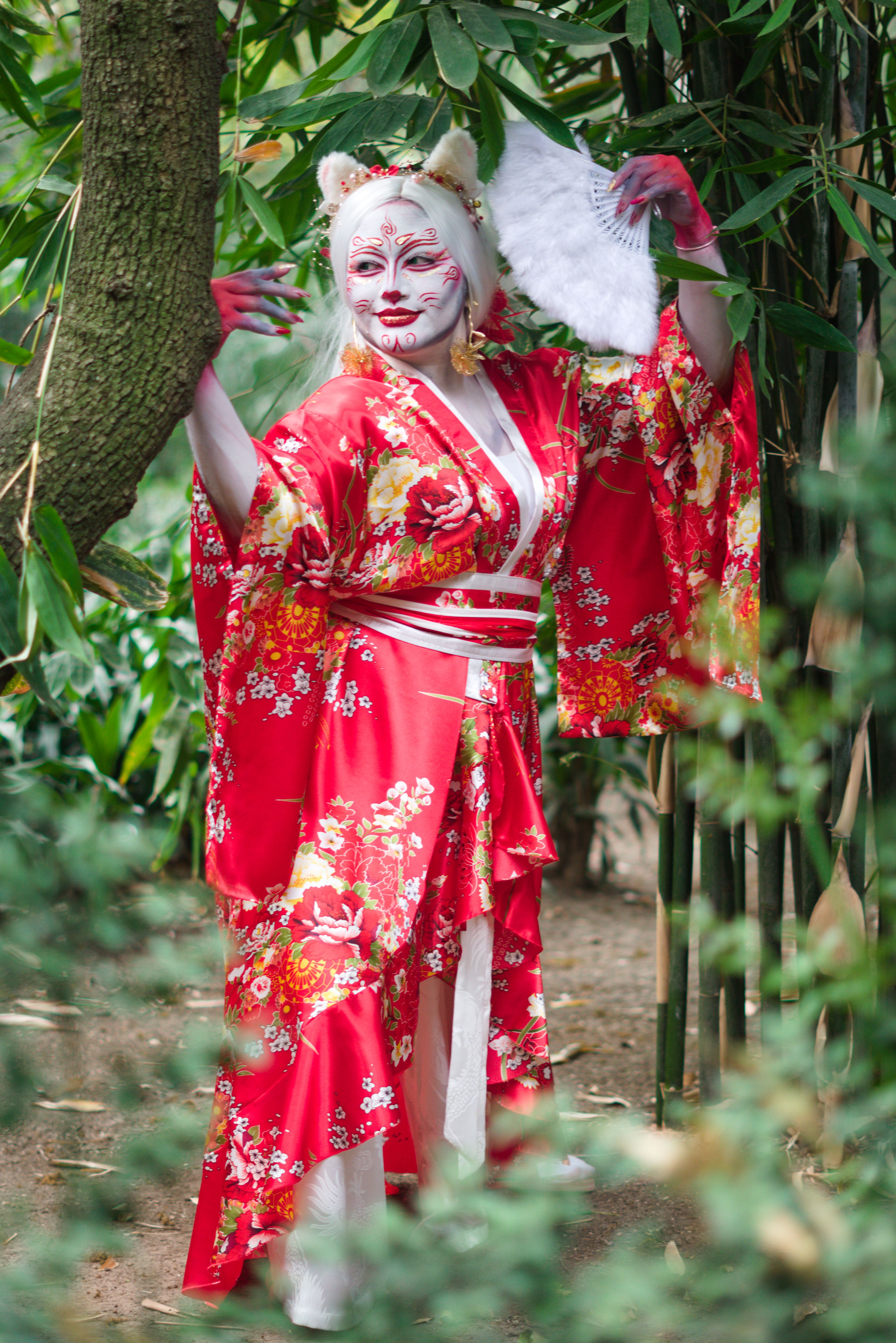 A cosplayer in a vibrant red floral kimono with fox makeup posing in a bamboo forest.