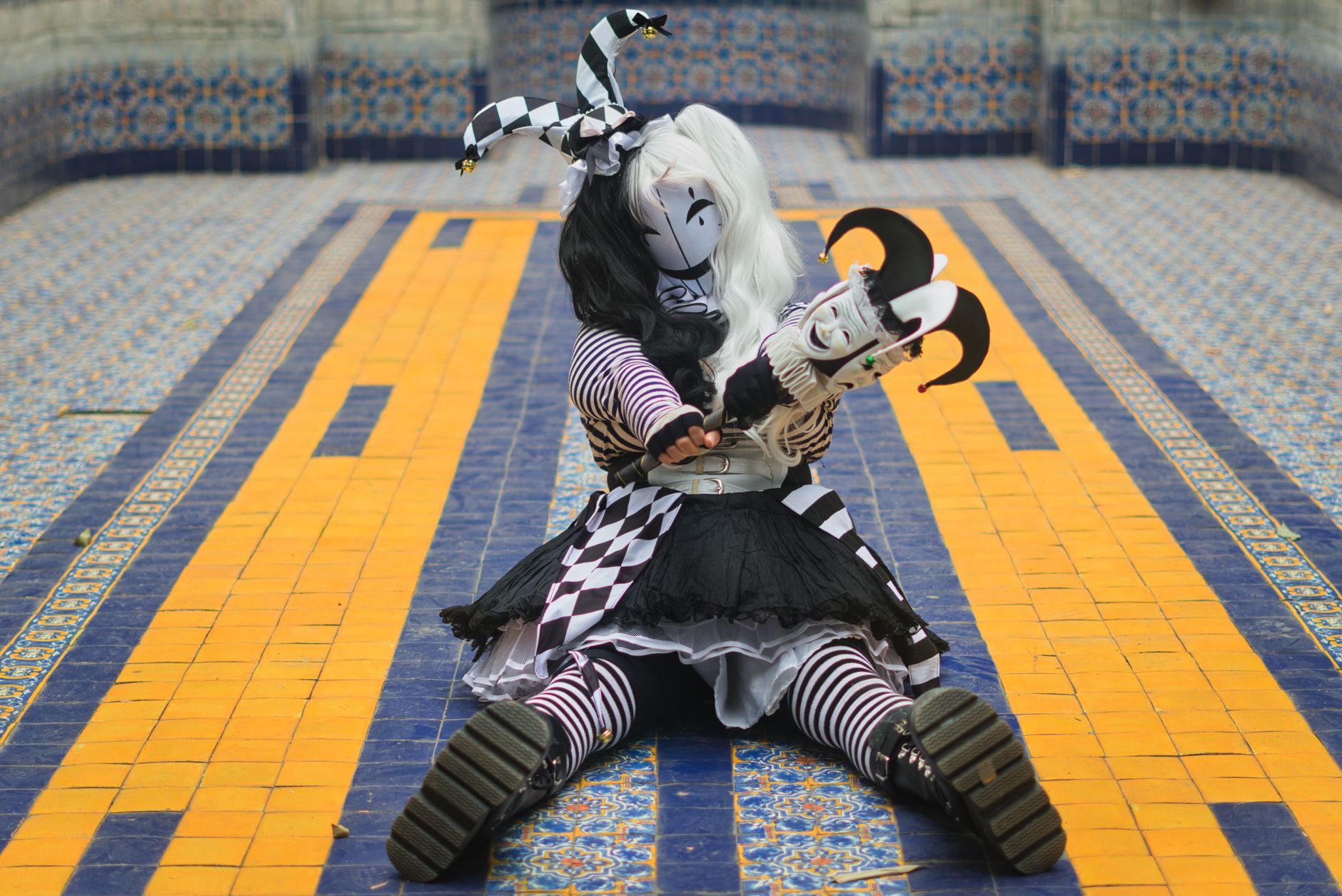 A harlequin dressed in black and white costume sits on a vibrant mosaic tiled floor.
