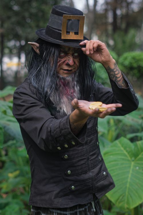 Free Halloween-themed image of a leprechaun holding gold coins in a lush outdoor setting. Stock Photo