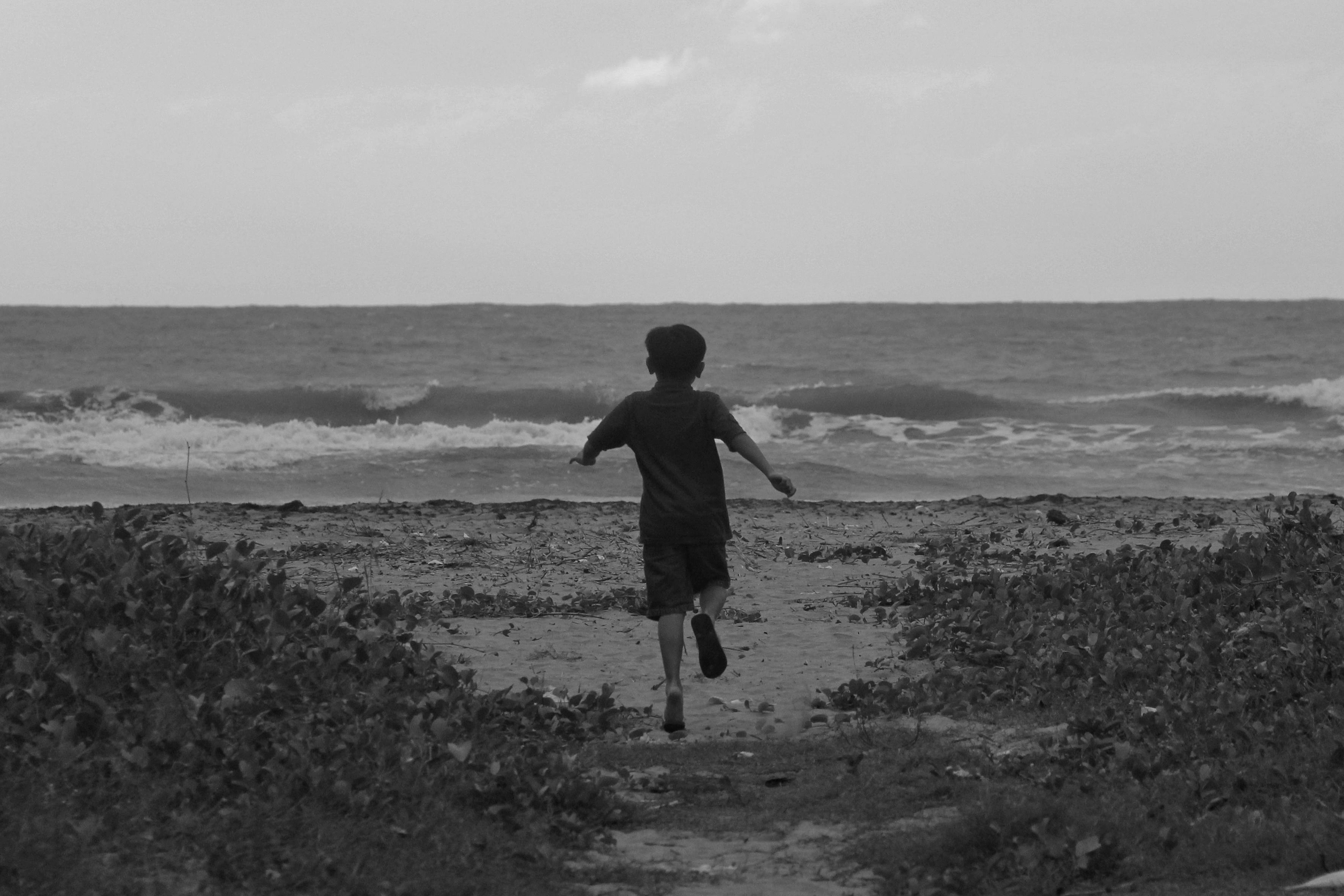 Gratis Un bambino corre gioioso verso l'oceano su una spiaggia sabbiosa, catturando un senso di libertà. Foto a disposizione